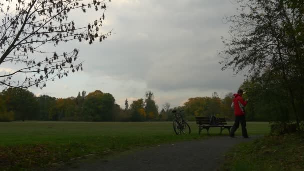 Турист Щелкает телефон в Parlk ходить скамейке Молодой человек смс Играя игры велосипед оставлен позади скамейки в облачно осенний парк день — стоковое видео