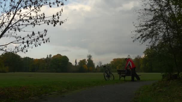 Rowerzysta turystycznych w ławce będzie urlop dostaje na rower i ruszanie w pochmurne jesienne dnia Park sportowca w czerwonej kurtce zielony trawnik chodnik — Wideo stockowe