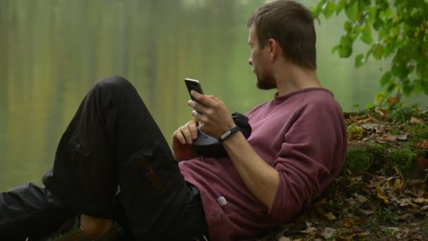 Touriste en lunettes 360Vr mensonges sur un sol Regarder la vidéo à l'aide de Smartphone Jeux Virtuels Clicks l'homme de dispositif dans le parc d'automne nuageux Jour par l'eau — Video
