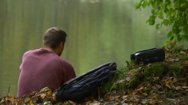 男人是谈话由电话坐在地面户外旅游是看视频 360 度玩游戏在公园由水地面 360vr 眼镜左边 — 图库视频影像