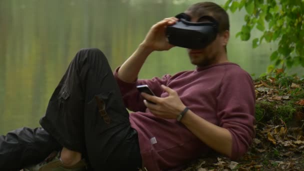 Turystycznych jest oglądania vr wideo w 360vr okulary na zewnątrz leży na ziemi w pochmurno Autumn dzień Park przez człowieka, woda jest, gry wirtualne gry odpoczynku — Wideo stockowe