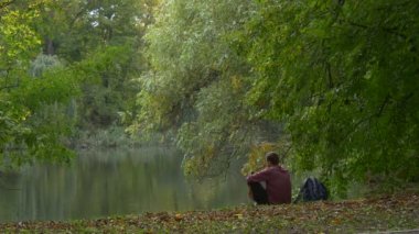 Adam Backpacker Sittng Gölü banka Park orman da yumuşak su dinlenme açık havada kuru yaprakları üzerinde bir yere yeşil ağaçlar sonbahar manzara, Looking