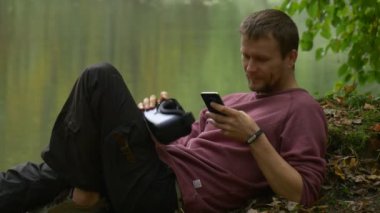 Yetişkin bir adam parkta gölün yanında akıllı telefon ve video kamera kullanıyor.