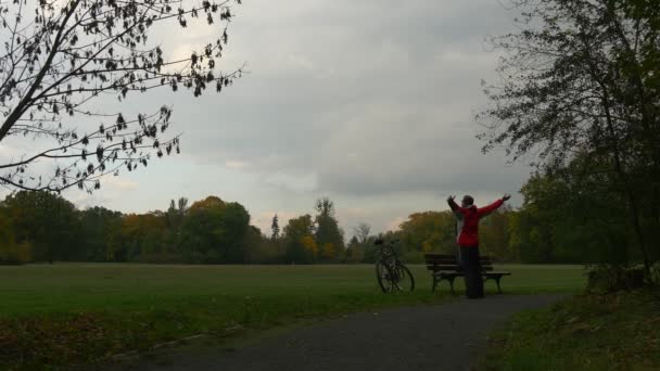 Człowiek stojący na ławce w parku wyciągnął zestaw w dół na ławeczce, drewniane, odpoczynek w Nature, rozglądając się na jesień krajobraz rowerów na ławce — Wideo stockowe