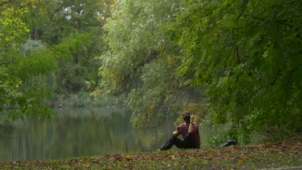Mann sitzt auf dem Boden in 360vr Helm Herbst Landschaft Tourist Video 360 Grad spielen virtuelle Spiele bewölkten Tag Park am Wasser — Stockvideo