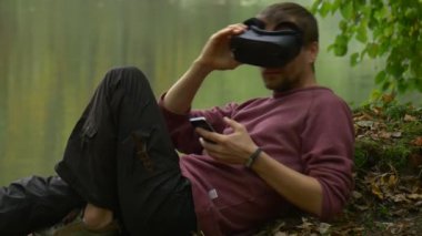 Yetişkin adam parkta gölün yanında VR gözlük ve akıllı telefon kullanıyor.
