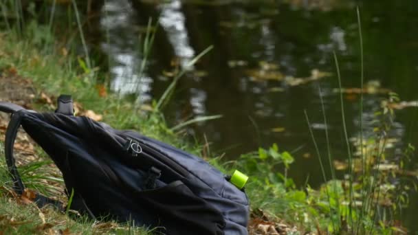 Toeristische links zwarte rugzak op de grond gras zonnige dag Park aan het Water droog gele bladeren drijvend door waterige oppervlak licht reflectoren op een rugzak — Stockvideo