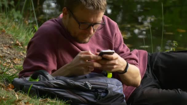 Tourist Clicks Mobile Phone Watching Video Playing Games Texting Clicks the Smartphone Lying on the Ground Man in Glasses in Sunny Day Park by the Water — Stok Video