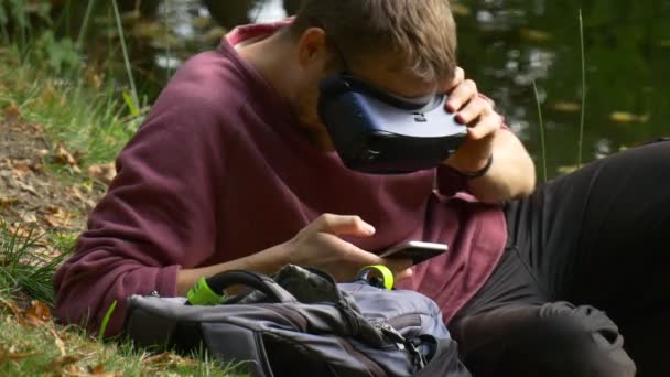 Muž v 360vr brýle sledování Video pomocí telefon venku hrát virtuální hry má vr Headset Off a brýle odpočívá v Sunny Park den u vody — Stock video