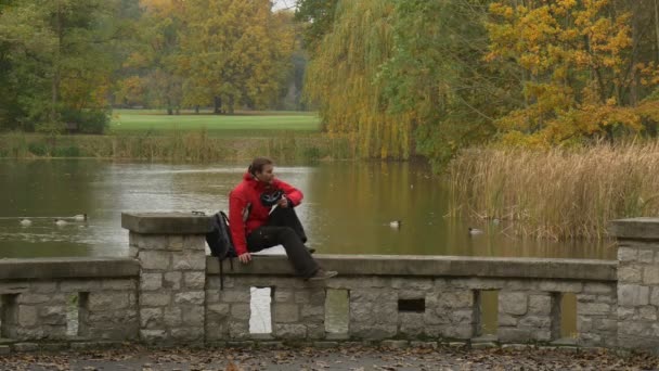 Turista v červené barvě drží 360vr brýle na mostě hraní her odpočívá v zatažené den parku na podzim při pohledu na vodu a kachny sledovat Video o 360 stupňů — Stock video