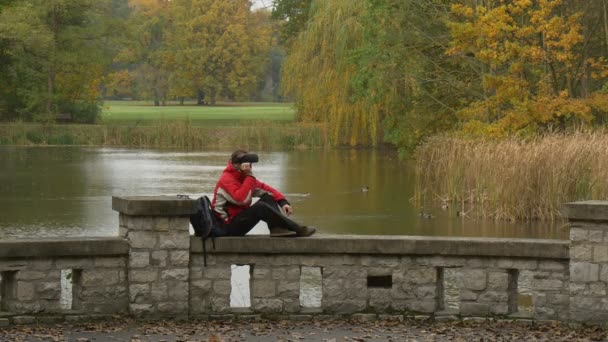 孤独在 vr 眼镜 Sittning 桥围栏看视频 360 度玩虚拟游戏在阴天在河或湖秋天公园旅游 — 图库视频影像