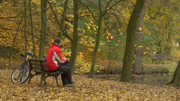 Ciclista viene collocato la sua moto si siede a panchina autunno giorno l'uomo sta riposando tra gli alberi d'oro nel parco terra è ricoperta di foglie gialle sentiero vicolo — Video Stock