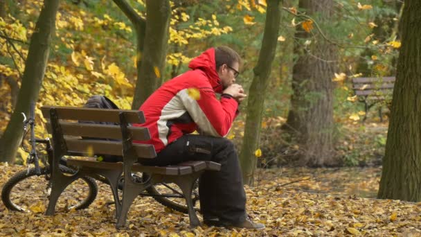 Le touriste se reposant sur un banc traverse ses jambes Le cycliste du jour d'automne est un arbre doré dans le parc Le vélo est laissé derrière le banc Les feuilles jaunes tombent — Video