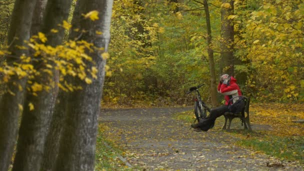 Solitaire touriste est assis sur le banc Journée d'automne Cycliste se repose parmi les arbres dorés dans le parc met sur les oreillettes obtient sol froid est recouvert de feuilles jaunes — Video