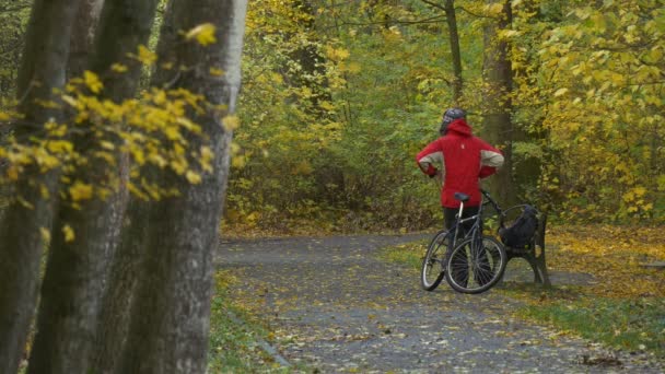 Turista toma la mochila fuera de caminatas por el parque Bicicleta en el banco en callejón Día de otoño ciclista está descansando entre los árboles de oro hojas amarillas en el suelo — Vídeo de stock