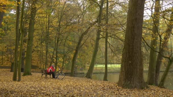 Man Sitting on a Bench Takes Earflaps Hat Off Resting Looking at Lake River Autumn Day Ciclista passa o tempo entre árvores douradas no parque folhas amarelas — Vídeo de Stock