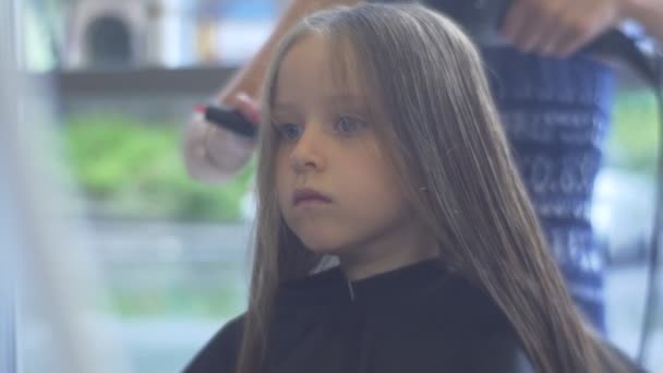 Friseur kämmt und trocknet blonde Kinderhaare Friseursalon Stylist macht eine Frisur kleines Mädchen sitzt mit ernstem Gesicht in peignoir — Stockvideo