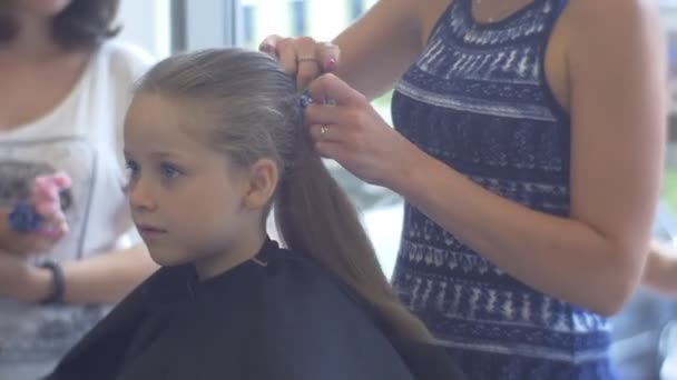 Стилист делает хвостик для блондинки клиентка ребенок в парикмахерской стилист делает прическу для серьезной маленькой девочки, сидя в черном Peignoir — стоковое видео