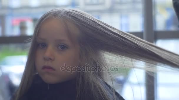 Beautician Dries Blonde Hairs Kid es un cliente Peluquería Estilista está haciendo un corte de pelo Niña está sentada en Peignoir Negro con cara seria — Vídeos de Stock