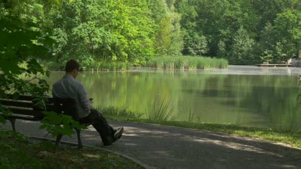 Homem usando telefone celular no parque descansando dia ensolarado em um banco junto ao Lago Turista está escrevendo leitura usando Internet Fresh Green Trees Reed Primavera Verão — Vídeo de Stock