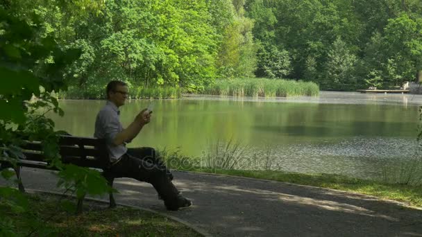 Homme avec tablette virtuelle assis dans le parc Homme d'affaires sur un banc Forex Trading Paiement Commande en ligne sur Internet Touriste par le lac frais ensoleillé été — Video