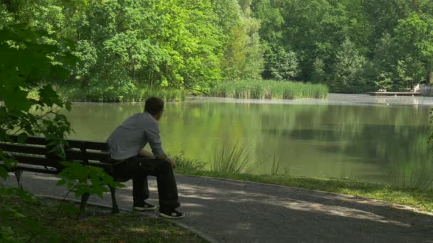 Homem descansando em um banco faz uma chamada no beco do parque telefônico junto ao lago Turista está falando Olhando para a água fresca árvores verdes suave água verão ensolarado — Vídeo de Stock