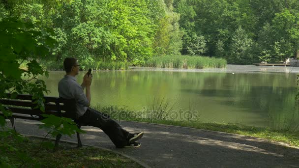 Tourist, der sich auf einer Bank ausruht, telefoniert auf der Parkallee am See Tourist spricht mit Blick auf das Wasser frische grüne Bäume glattes Wasser sonniger Sommer — Stockvideo