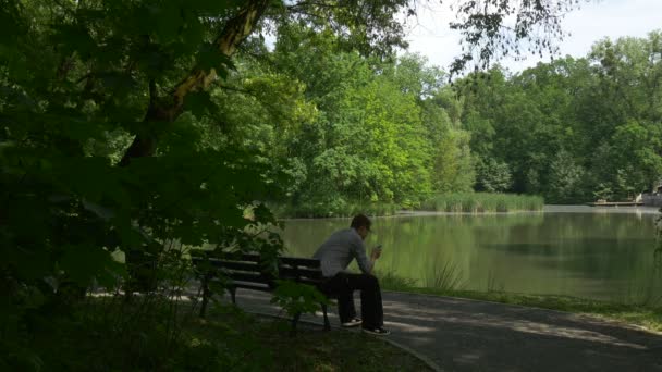 L'uomo fa una chiamata sul telefono cellulare in parco seduto su una panchina vicino al lago Turista sta messaggiando lettura utilizzando Internet alberi verdi freschi Sunny Summer — Video Stock