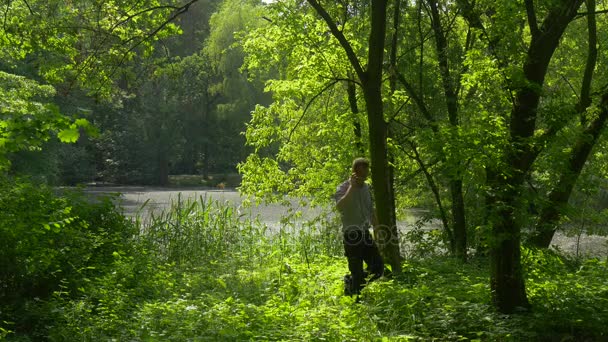 Homem se levanta Salta sobre a grama alta no parque Segurando telefone Descanso junto ao lago na floresta Frescura Árvores verdes Sunny Spring Day Luz solar vem através de filiais — Vídeo de Stock