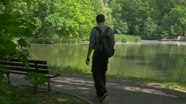 Ο άνθρωπος έρχεται στο υπόλοιπο στο πάρκο στέκεται στο πάγκο από τουριστική λίμνη ανάμεσα σε φρέσκα πράσινα δέντρα ηλιόλουστη, ανοιξιάτικη ημέρα ομαλή νερό ασφαλτοστρωμένο μονοπάτι ξύλινο πάγκο — Αρχείο Βίντεο
