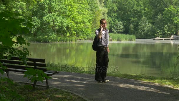 Backpacker делает звонок на мобильный телефон, стоящий в Аллее Прогулка по парку Отдых на гладком озере Турист в парке Свежие зеленые деревья Солнечный летний день — стоковое видео