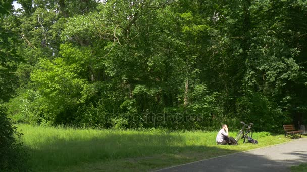 Homem ciclista sentado no parque e conversando por telefone Passe o tempo na natureza Árvores verdes frescas Dia ensolarado de verão Banco de madeira no beco Turista está descansando — Vídeo de Stock