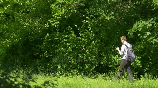 Homem Clicks Telefone Caminhando no Parque Segurando Livro Turista Passe o tempo na natureza Entre árvores verdes frescas e grama ensolarado Verão Dia Texting Usando o Dispositivo — Vídeo de Stock