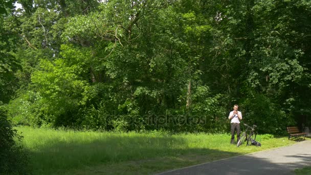 Touriste cycliste assis dans le parc Clicks Téléphone Passez du temps à la nature Arbres verts frais Ensoleillé Journée d'été Banc en bois dans l'allée Touriste se repose — Video