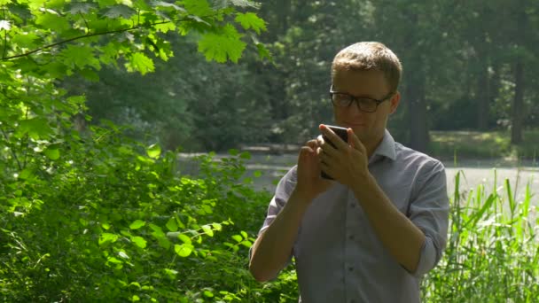 Jonge Man in groene weelderige struiken maakt een gesprek praten over telefoon rusten in Park frisheid van de groene bomen in de zonnige zomer zonlicht Man besteedt weekend landschap — Stockvideo