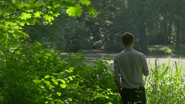 Tourist is Texting on Phone Near Lake Standing Among Green Lush Bushes Sunlight Cyclists Are Riding on the Other Side Man Spends Weekends in Spring — Stock Video