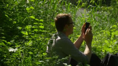 Bir yere Park yeşil yemyeşil çim çalıların genç adam Park tazelik güneş ışığı erkekte dinlenme içinde oturan turist tıklama telefon ilkbahar sonlarında harcıyor