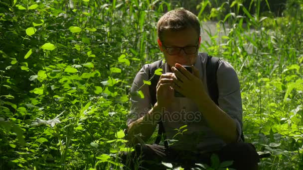 Turystycznych, wywołanie na telefon siedzi wśród trawy odpoczynku w światło słoneczne świeżość Park zielony bujnej trawy krzewy człowiek spędza weekendy na wiosnę za pomocą cyfrowego urządzenia — Wideo stockowe