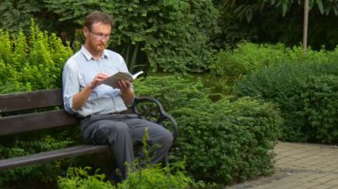 Elderly Man With Glasses Reading a Book on a Park Bench. Then Put the Book Aside and Looking Thoughtful. Green Trees and Bushes Around the Bench. Summer Day