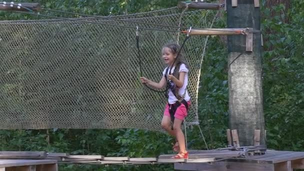 Happy Girl è nel parco avventura Passeggiando vicino al ponte di corda durante la giornata estiva Il bambino si diverte occupato da Sport Kid è fissato ai cavi con l'attrezzatura da arrampicata Fissa — Video Stock