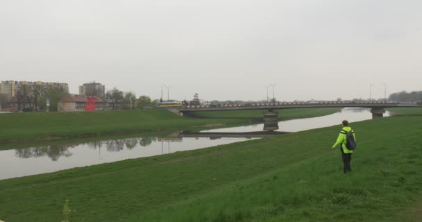 Mochilero en el campo verde patea la hierba Absently Descanso en el paisaje Césped verde Paisaje urbano en un puente de coche Horizonte sobre el río Nublado Primavera de Europa — Vídeos de Stock