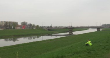 Turist nehir manzara yeşil çim Cityscape, yerdeki bir ufuk araba Köprüsü üzerinden River bulutlu gün adam alır üzerinde oturuyor ve yaprakları