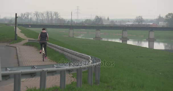 Mujer está montando una bicicleta a lo largo del paisaje del río Green Lawn Valley Cityscape Car Bridge Over Smooth River Vehicles Are Driven in Cloudy Spring Day — Vídeo de stock
