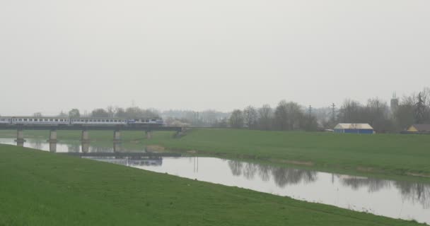 Passenger Train is Moving Over River by Bridge Rural Landscape Green Lawn Reflection in Smooth Water Small Village Houses Foggy Cloudy Spring Day — Stock Video