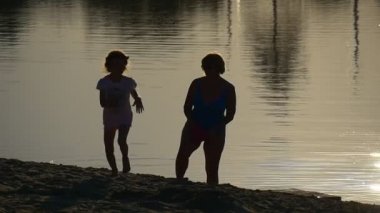 Çocuk ve kadın siluetleri nehir Sandy Beach küçük kız, çalışan tarafından kıyı şeridi anne büyükanne olduğunu yürüyüş arkasında kızı günbatımı yaz