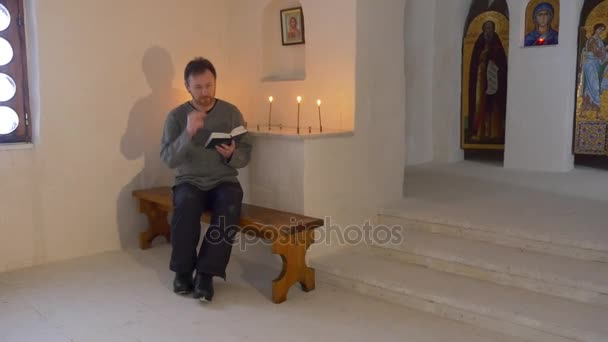 Χριστούγεννα στο Svyatogorsk μοναστήρι άνδρας διαβάζει την Αγία Γραφή υπό το πρίσμα εκκλησία κεριά εικόνες εικονίδια βρίσκονται σε ένα τοίχο λάτρη κάθεται στον πάγκο — Αρχείο Βίντεο