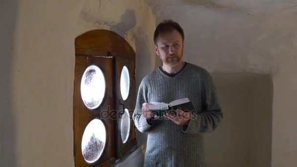 Homem com Bíblia no quarto pequeno no mosteiro adorador é leitura atenta Lattice porta turístico na capela dentro o giz caverna Tour para Svyatogorsk — Vídeo de Stock