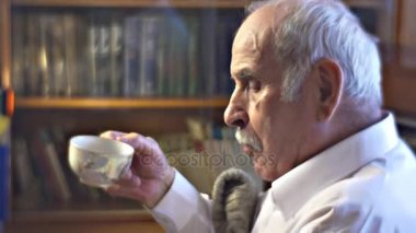 Yaşlı adam yeme ve içme çay Cosy oda iç soğuk kış günü evde memnuniyet dedesi Beyaz gömlekli rahat koltukta oturuyor