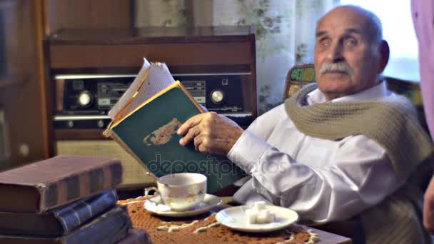 Сын поговорить со старым отцом, который читает старую книгу История жизни старика старший человек выпил чай и наслаждается своими воспоминаниями груду книг на столе — стоковое видео