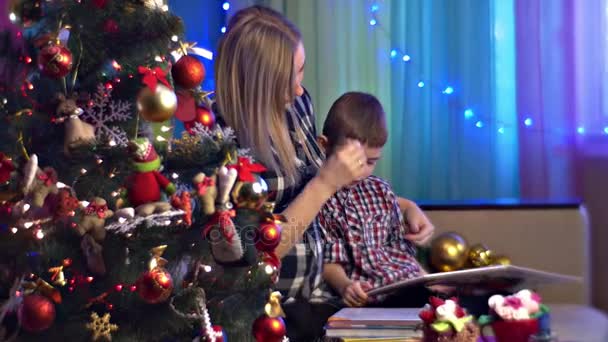 Genç annedir okuma kitaplar ile onun oğlu aile kutluyor Noel dekore edilmiş yeni yıl ağacı hamile anne ve küçük çocuğu bekleyen için bebek rahat numaralı oda — Stok video